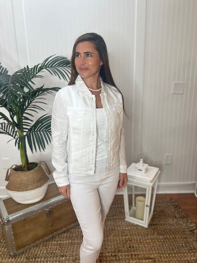A woman stands wearing a white linen jacket with a pearl necklace, white tank top and white pants with plant and antique trunk in the background