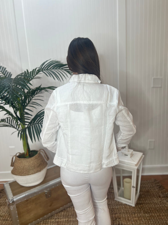 A woman stands wearing a white linen jacket with white pants with plant and antique trunk in the background
