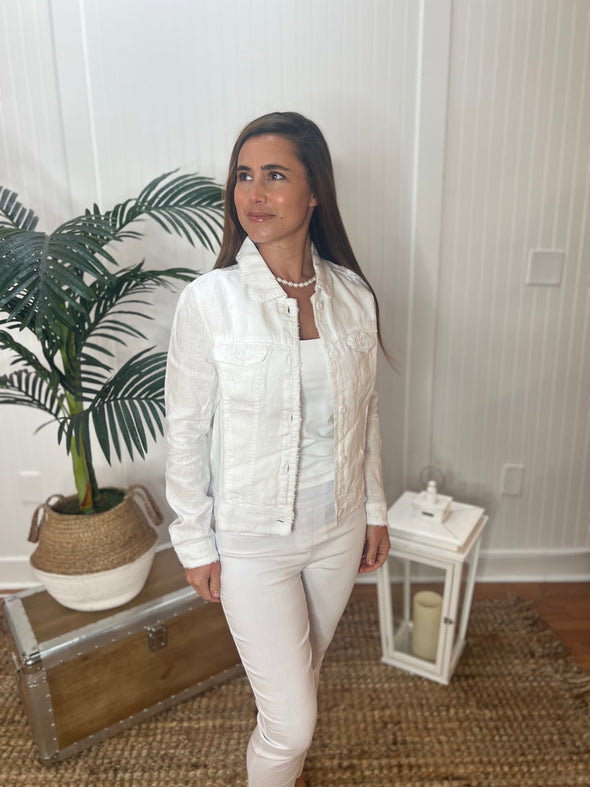 A woman stands wearing a bright white jacket with a pearl necklace, white tank top and white pants with plant and antique trunk in the background