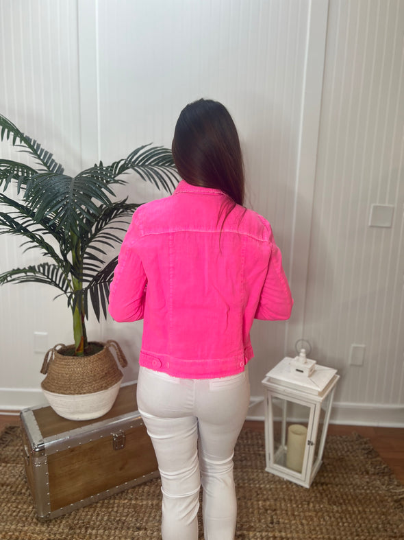 A woman stands facing away from camera wearing hot pink jacket and white pants with plant and antique trunk in the background