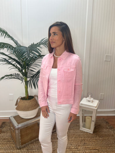 A woman stands angled wearing a light pink linen jacket with a white tank topand white pants with plant and antique trunk in the background