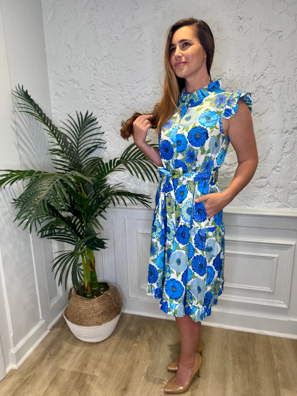 Smiling model in front of a white wall with a plant in a corner wearing the Patty Kim Megan Dress - Blue Zinnias. A midi length flutter sleeve and collar, with a tie waist and a ruffle at the bottom hem, with blue and green flower pattern
