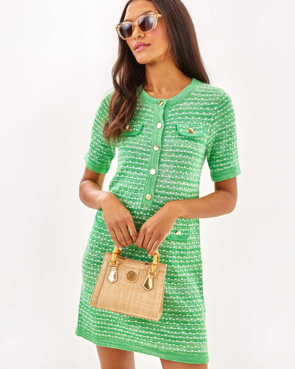 Model in the Lilly Pulitzer Theodorah Sweater Dress - Fauna Green Metallic Marl holding a woven purse. A mini knitted dress with gold buttons down front and on front pockets. 