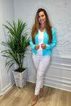 Smiling model in front of a white wall with a green plant in the corner. She's wearing the Lulu B Linen Travel Jacket - Seafoam, a light linen jacket with 2 front pockets and lightly frayed hems. Paired with a white shirt and white pants. 