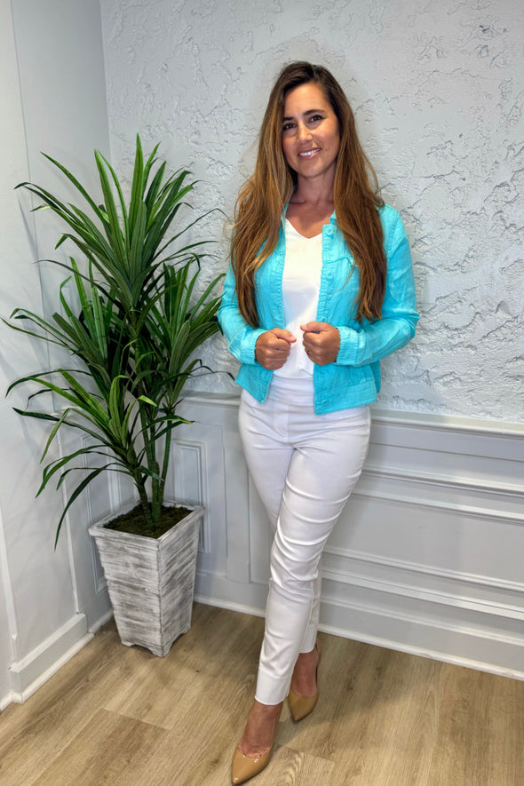 Smiling model in front of a white wall with a green plant in the corner. She's wearing the Lulu B Linen Travel Jacket - Seafoam, a light linen jacket with 2 front pockets and lightly frayed hems. Paired with a white shirt and white pants. 