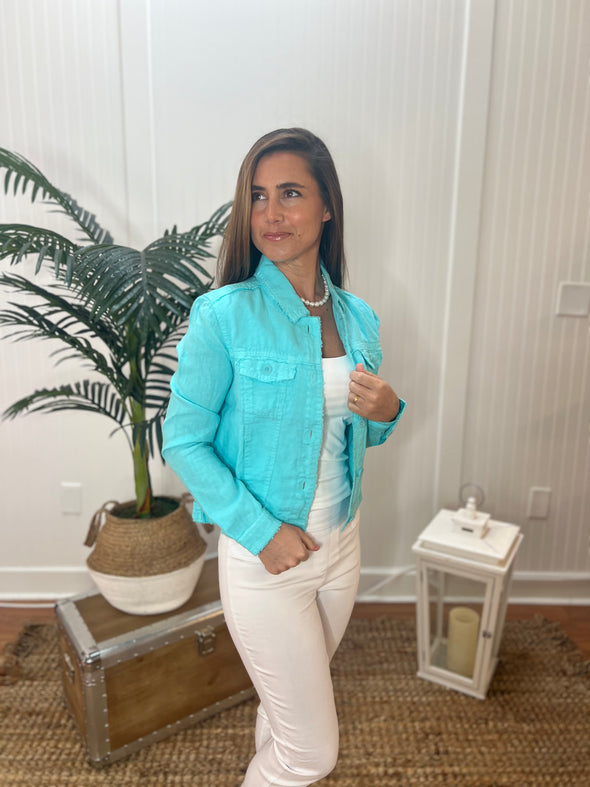 A woman stands wearing a bright aqua linen jacket with a pearl necklace, white tank top and white pants with plant and antique trunk in the background