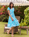 Smiling model outside in front of a char, side table, and umbrella, twirling in the Jude Connally Libby Dress - Enchanted Sea Aqua
