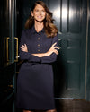 Model in the Jude Connally Midge Dress - Navy/Gold. A navy dress with gold buttons down the front, a shirt style collar, and split arm hems, standing in front of a dark green door