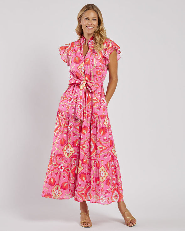 Smiling model in the Jude Connally Mirabella Dress in Gilded Garden Peony Pink Gold. A pink orange and gold midi dress with flutter sleeves and tie belt with a floral/geometric pattern