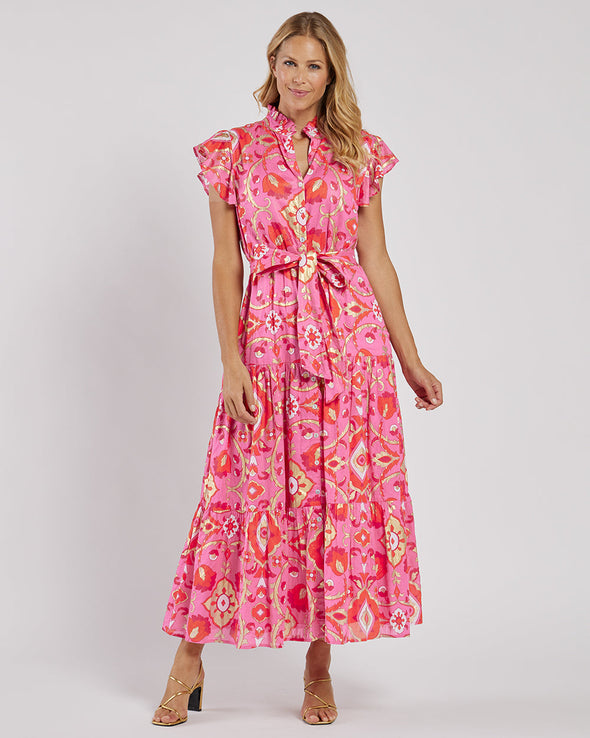 Smiling model in the Jude Connally Mirabella Dress in Gilded Garden Peony Pink Gold. A pink orange and gold midi dress with flutter sleeves and tie belt with a floral/geometric pattern