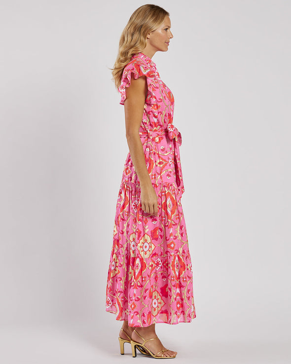 Side view of the Jude Connally Mirabella Dress in Gilded Garden Peony Pink Gold. A pink orange and gold midi dress with flutter sleeves and tie belt with a floral/geometric pattern