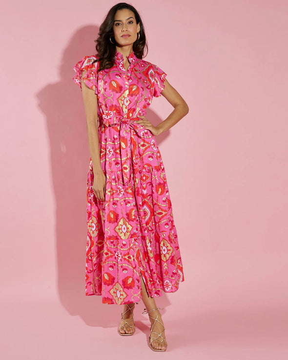 Model in front of a pink wall wearing the Jude Connally Mirabella Dress in Gilded Garden Peony Pink Gold. A pink orange and gold midi dress with flutter sleeves and tie belt with a floral/geometric pattern