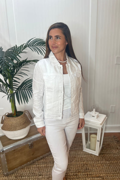 A woman stands wearing a white linen jacket with a pearl necklace, white tank top and white pants with plant and antique trunk in the background