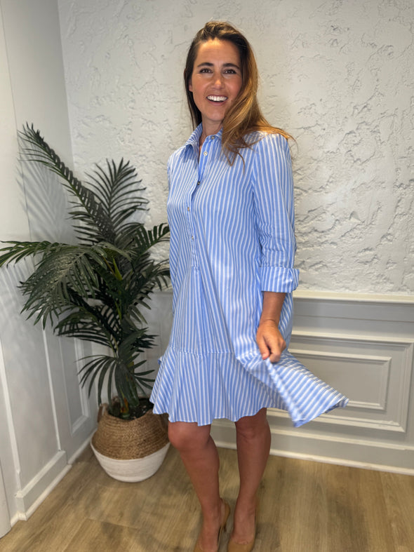 Smiling model in front of a white wall and a tropical plant in the corner wearing the Duffield Lane Iris Drop Waist Shirt Dress - Light Blue Double Stripe. A mini/knee length, long sleeve, seersucker dress. It has a collar and buttons down the front and a ruffle at the bottom hem. 