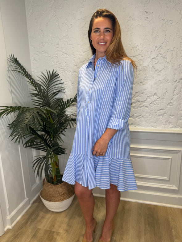 Smiling model in front of a white wall and a tropical plant in the corner wearing the Duffield Lane Iris Drop Waist Shirt Dress - Light Blue Double Stripe. A mini/knee length, long sleeve, seersucker dress. It has a collar and buttons down the front and a ruffle at the bottom hem. 