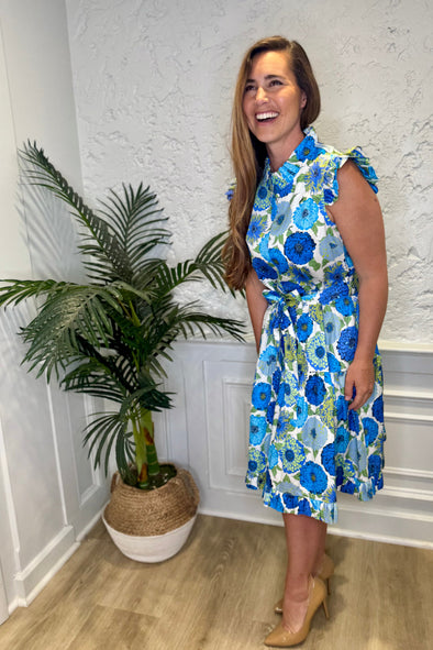 Smiling model in front of a white wall with a plant in a corner wearing the Patty Kim Megan Dress - Blue Zinnias. A midi length flutter sleeve and collar, with a tie waist and a ruffle at the bottom hem, with blue and green flower pattern