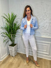 Smiling model in front of a white wall with a green plant in the corner. She's wearing the Lulu B Linen Travel Jacket - Seersucker, a light linen jacket with 2 front pockets and lightly frayed hems. Paired with a white shirt and white pants. 