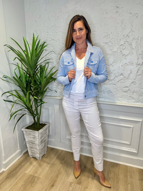 Smiling model in front of a white wall with a green plant in the corner. She's wearing the Lulu B Linen Travel Jacket - Seersucker, a light linen jacket with 2 front pockets and lightly frayed hems. Paired with a white shirt and white pants. 