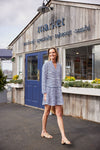 Smiling model in front of a market wearing a mini length, long sleeved, tiered dress with navy and white stripes. She is also wearing nude sandals and carrying a woven bag
