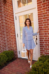 Smiling model walking out of a door or a brick building onto a brick path. She is wearing a mini length, long sleeved, tiered dress with navy and white stripes. She is also wearing white sandals and carrying a white and woven bag. 