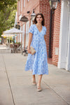 Woman in a blue floral dress standing on a sidewalk with a brick building and outdoor seating area in the background.
