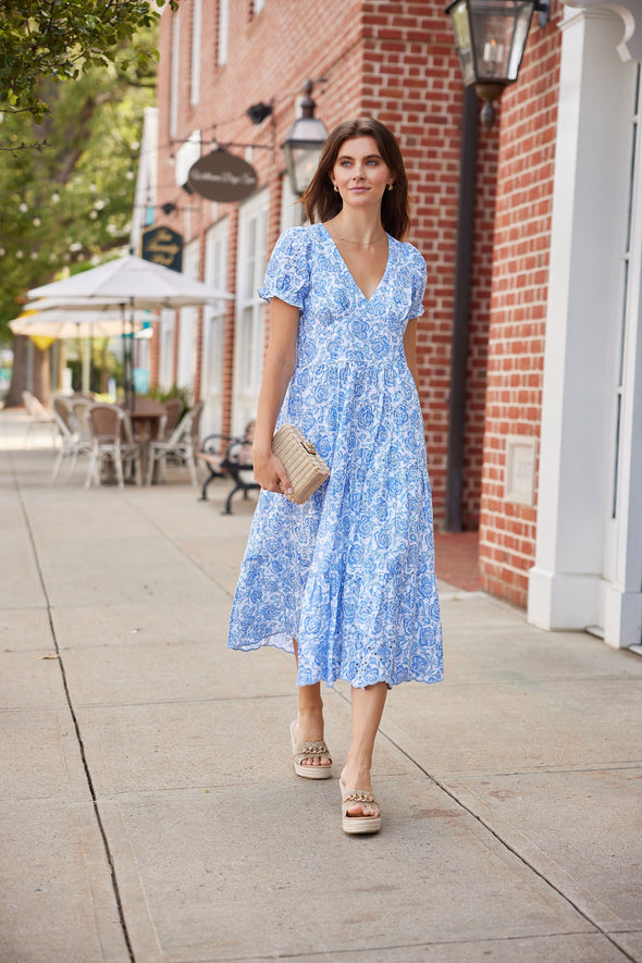 Woman in a blue floral dress standing on a sidewalk with a brick building and outdoor seating area in the background.