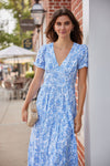 Woman in a blue floral dress standing against a wall outdoors.