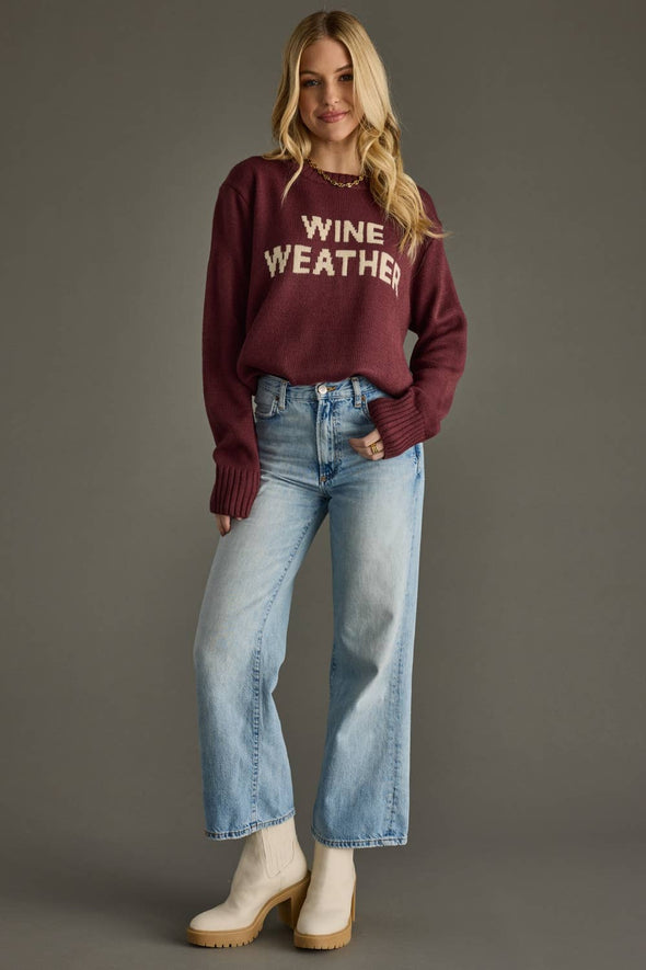 Woman wearing a maroon sweater with 'Wine Weather' text and blue jeans on a gray background