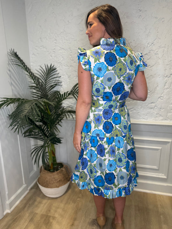 Back view of model in front of a white wall with a plant in a corner wearing the Patty Kim Megan Dress - Blue Zinnias. A midi length flutter sleeve and collar, with a tie waist and a ruffle at the bottom hem, with blue and green flower pattern