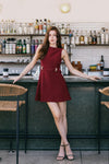 Model in a sleeveless red dress with a boatneck neckline a belt with gold detailing standing in a bar setting