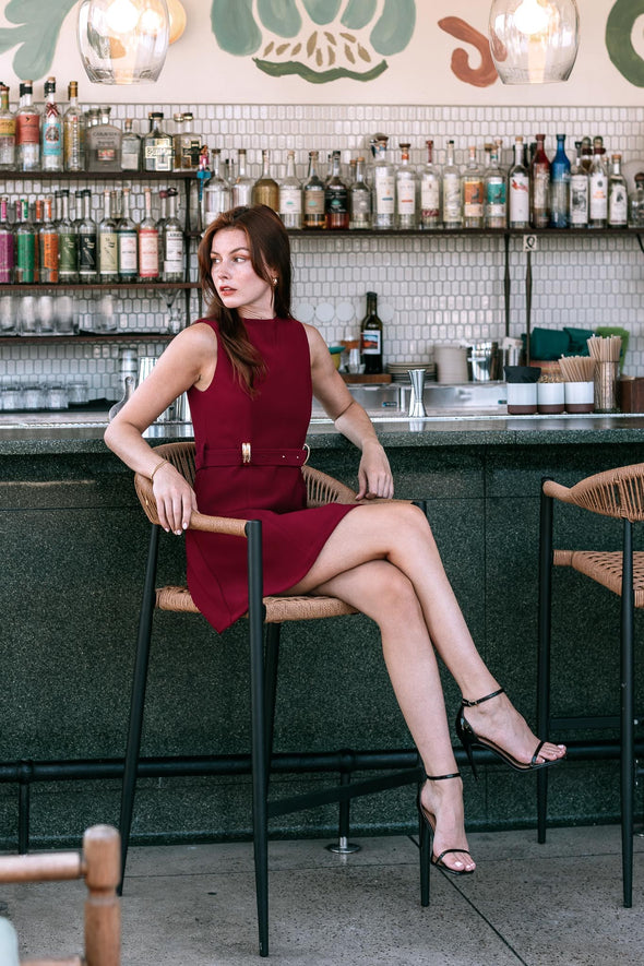 Model in a sleeveless red dress with a boatneck neckline a belt with gold detailing sitting in a bar chair at a bar