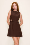 Model in a sleeveless brown dress with a boatneck neckline a belt with gold detailing standing in a bar setting