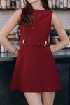 Model in a sleeveless red dress with a boatneck neckline a belt with gold detailing standing in a bar setting