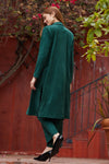 Woman wearing a green coat and black shoes against a red wall with plants.