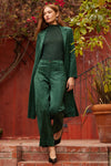 Woman in a green suede outfit standing on steps with a red wall and plants in the background