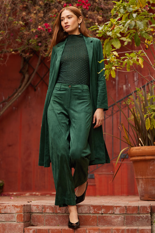 Woman in a green suede outfit standing on steps with a red wall and plants in the background