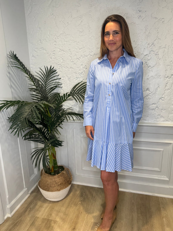 Smiling model in front of a white wall and a tropical plant in the corner wearing the Duffield Lane Iris Drop Waist Shirt Dress - Light Blue Double Stripe. A mini/knee length, long sleeve, seersucker dress. It has a collar and buttons down the front and a ruffle at the bottom hem. 