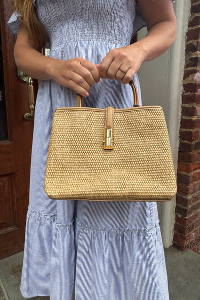 Model outside holding the Chatham Straw Bag - tan. A woven bag with tan trim and a bamboo handle 