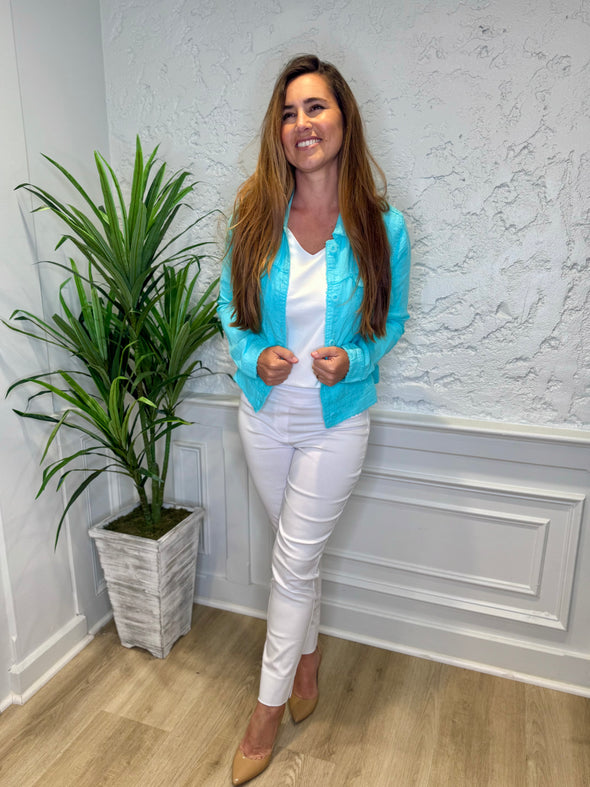 Smiling model in front of a white wall with a green plant in the corner. She's wearing the Lulu B Linen Travel Jacket - Seafoam, a light linen jacket with 2 front pockets and lightly frayed hems. Paired with a white shirt and white pants. 