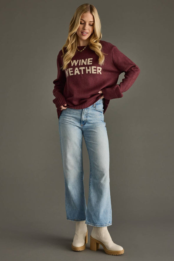 Woman wearing a maroon sweater with 'WINE WEATHER' text, blue jeans, and white boots on a gray background