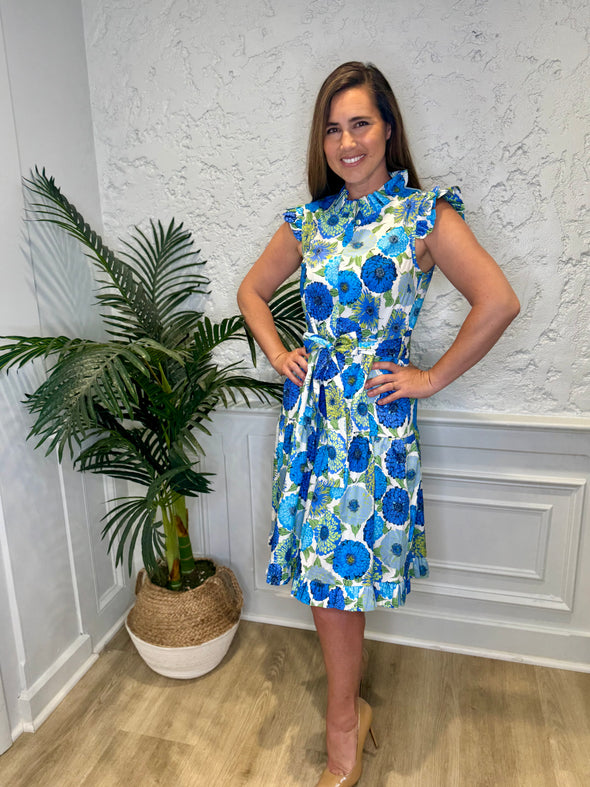 Smiling model with hands on hips in front of a white wall with a plant in a corner wearing the Patty Kim Megan Dress - Blue Zinnias. A midi length flutter sleeve and collar, with a tie waist and a ruffle at the bottom hem, with blue and green flower pattern