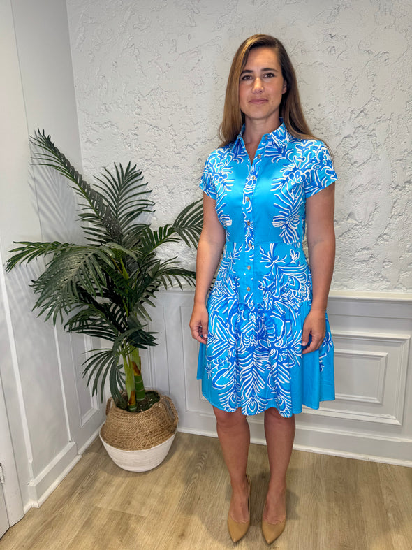Smiling model in front of a white wall with a plant in the corner, in the Patty Kim Colette Dress - Aqua Rococo. A short sleeved mini dress with a drop waist and buttons down the front. A white and royal blue floral pattern on an aqua background
