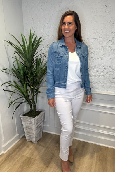 Smiling model in the Lulu B Denim Travel Jacket - Blue Jean in front of a white wall with a plant in the corner. A lightweight chambray jacket paired with a white shirt and white pants