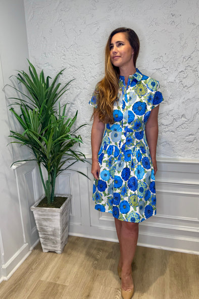 Model in front of a white wall in the Patty Kim Collette Dress - Blue Zinnias. A knee length dress with short sleeves and a drop waist and buttons down the front