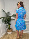 Back view of Smiling model in front of a white wall with a plant in the corner, in the Patty Kim Colette Dress - Aqua Rococo. A short sleeved mini dress with a drop waist and buttons down the front. A white and royal blue floral pattern on an aqua background