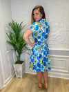 Back view of model in front of a white wall in the Patty Kim Collette Dress - Blue Zinnias. A knee length dress with short sleeves and a drop waist and buttons down the front