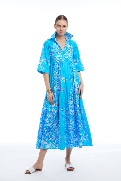 Woman wearing a bright blue maxi dress with white floral pattern. The dress has a V-neck, flared sleeves, and a tiered skirt. She’s wearing white sandals and standing against a plain white background.