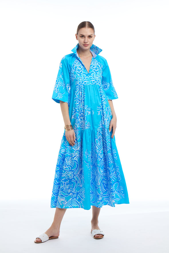 Woman wearing a bright blue maxi dress with white floral pattern. The dress has a V-neck, flared sleeves, and a tiered skirt. She’s wearing white sandals and standing against a plain white background.