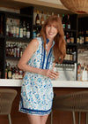 Smiling model at a bar holding a glass of champagne in the Cabana Life Bubbly Sleeveless Tunic Cover Up. A sleeveless mock neck shift dress with blue flowers on a white background and royal blue trim around the neckline and bottom hem