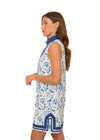 Model holding a glass of champagne in the Cabana Life Bubbly Sleeveless Tunic Cover Up. A sleeveless mock neck shift dress with blue flowers on a white background and royal blue trim around the neckline and bottom hem